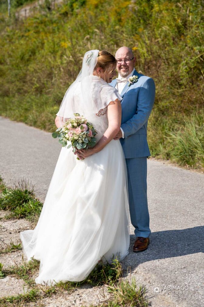 Bräutigam hält seine Braut in seinem Arm, Braut versteckt den Brautstrauß hinter ihrem Rücken, Hochzeitsfotografie, Paarshooting